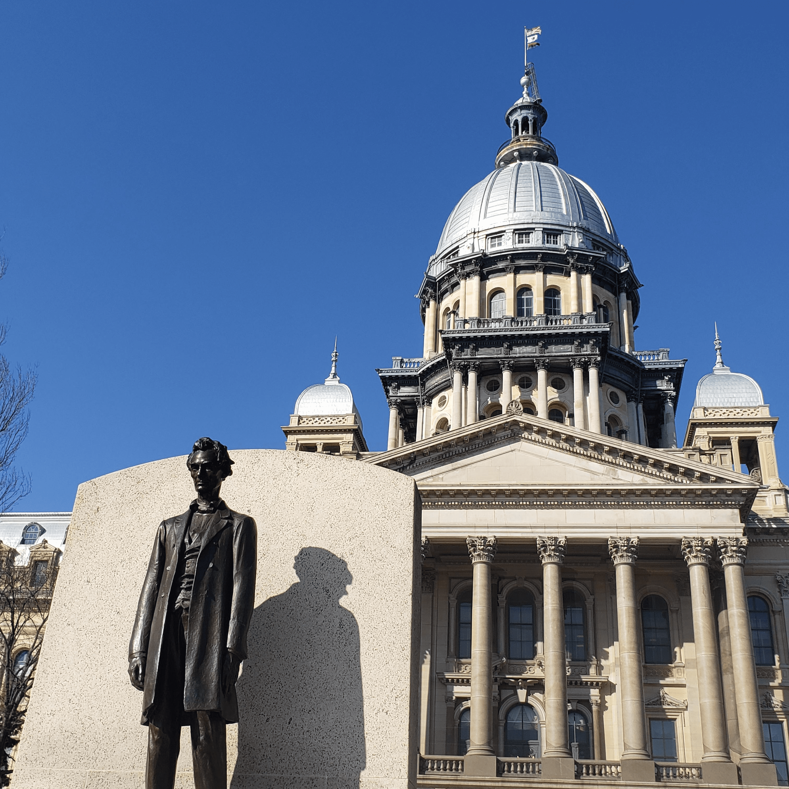 The statue of Abraham Lincoln in front of the IL State Capitol building and a blue sky.